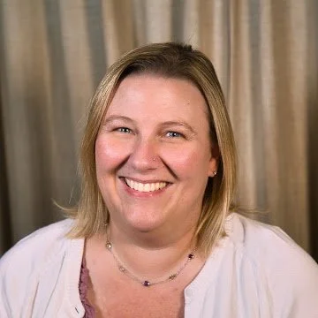A woman with blonde hair smiling in front of beige curtains.