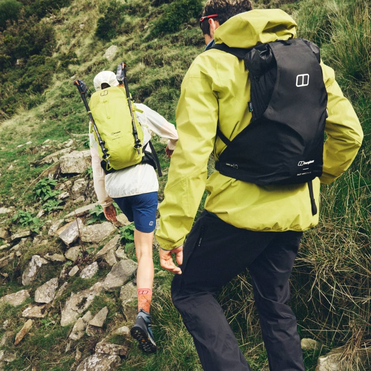 Two hikers walking up a rocky trail on a green hillside, wearing outdoor gear and backpacks.