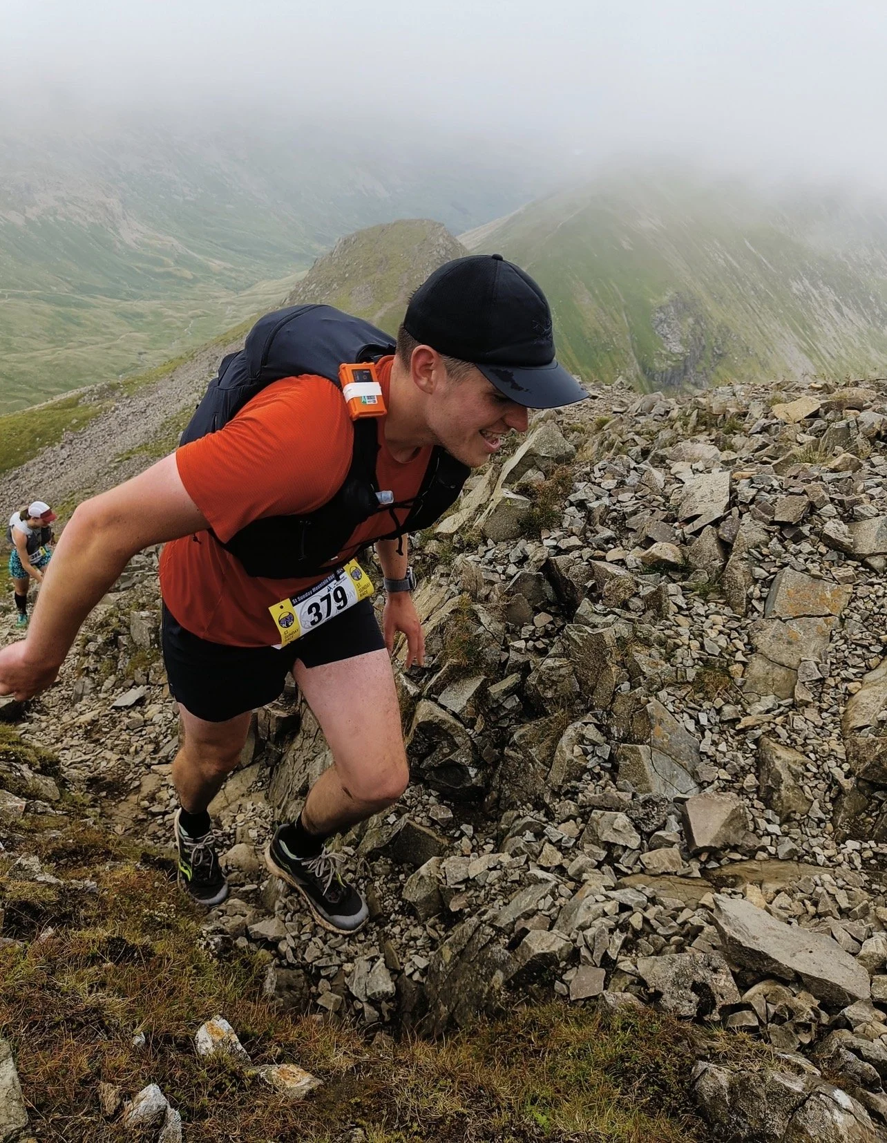 A man participating in a mountain trail running race, wearing a black cap, orange shirt, black shorts, and a backpack, climbing up a rocky trail with misty mountains in the background.