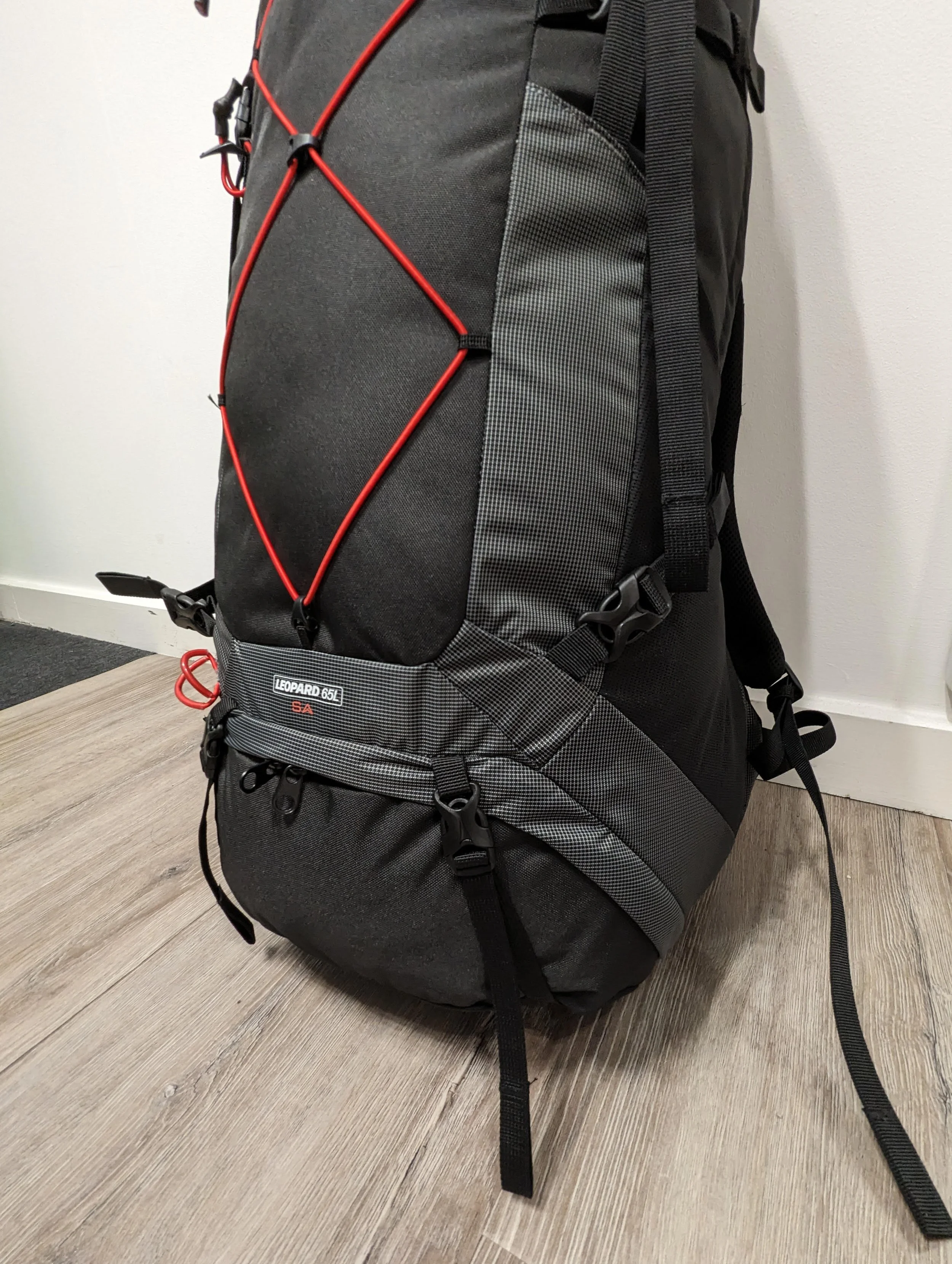 A black and gray hiking backpack with red bungee cords and straps, situated on a wooden floor against a plain white wall.