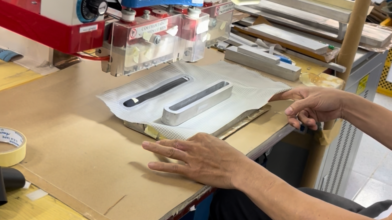 A person using a machine to shape or polish a long, flat metal object in a workshop or manufacturing setting.
