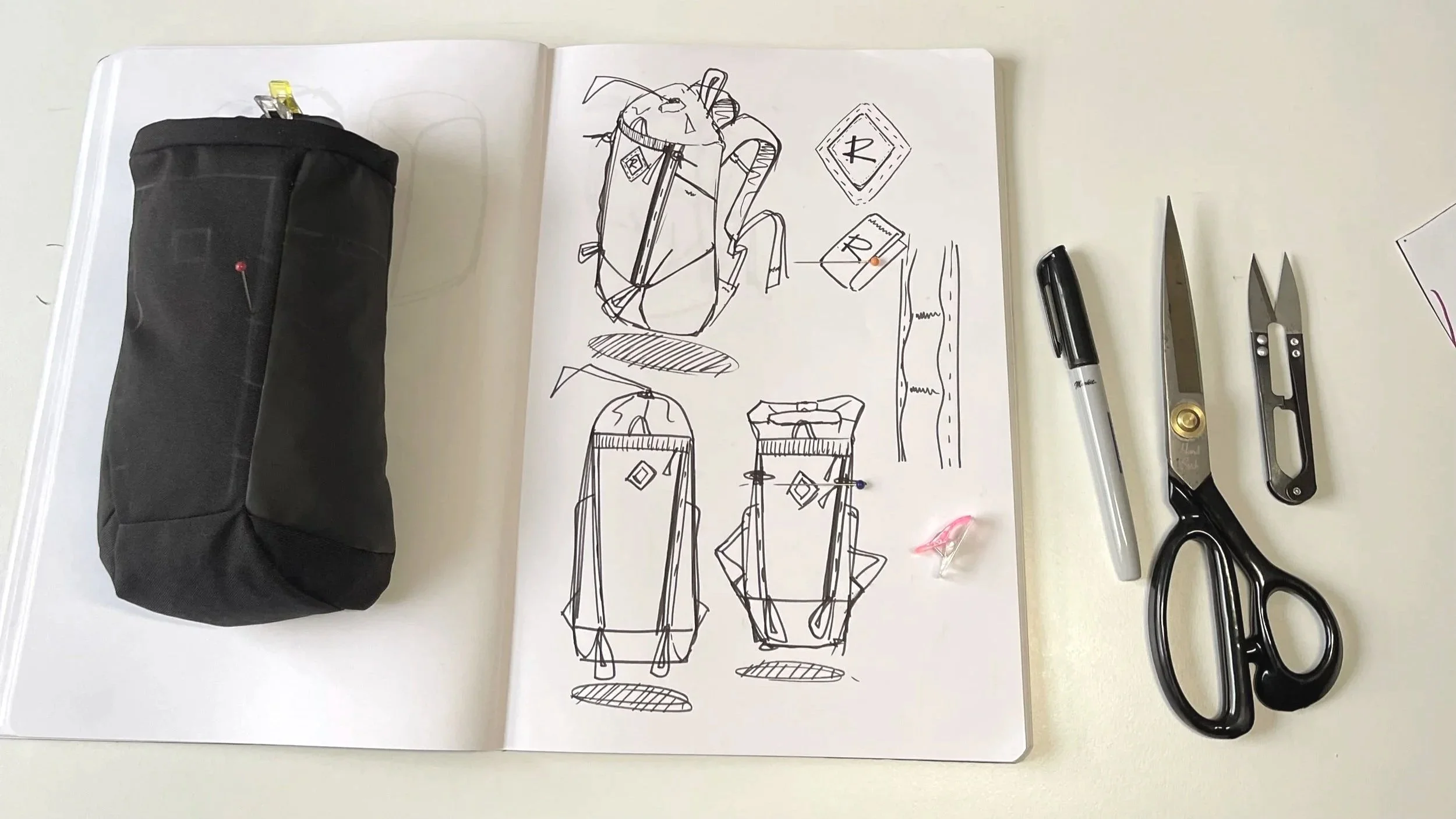 Sketches of backpack designs on an open notebook with a black pencil case, scissors, and a black marker on a white surface.