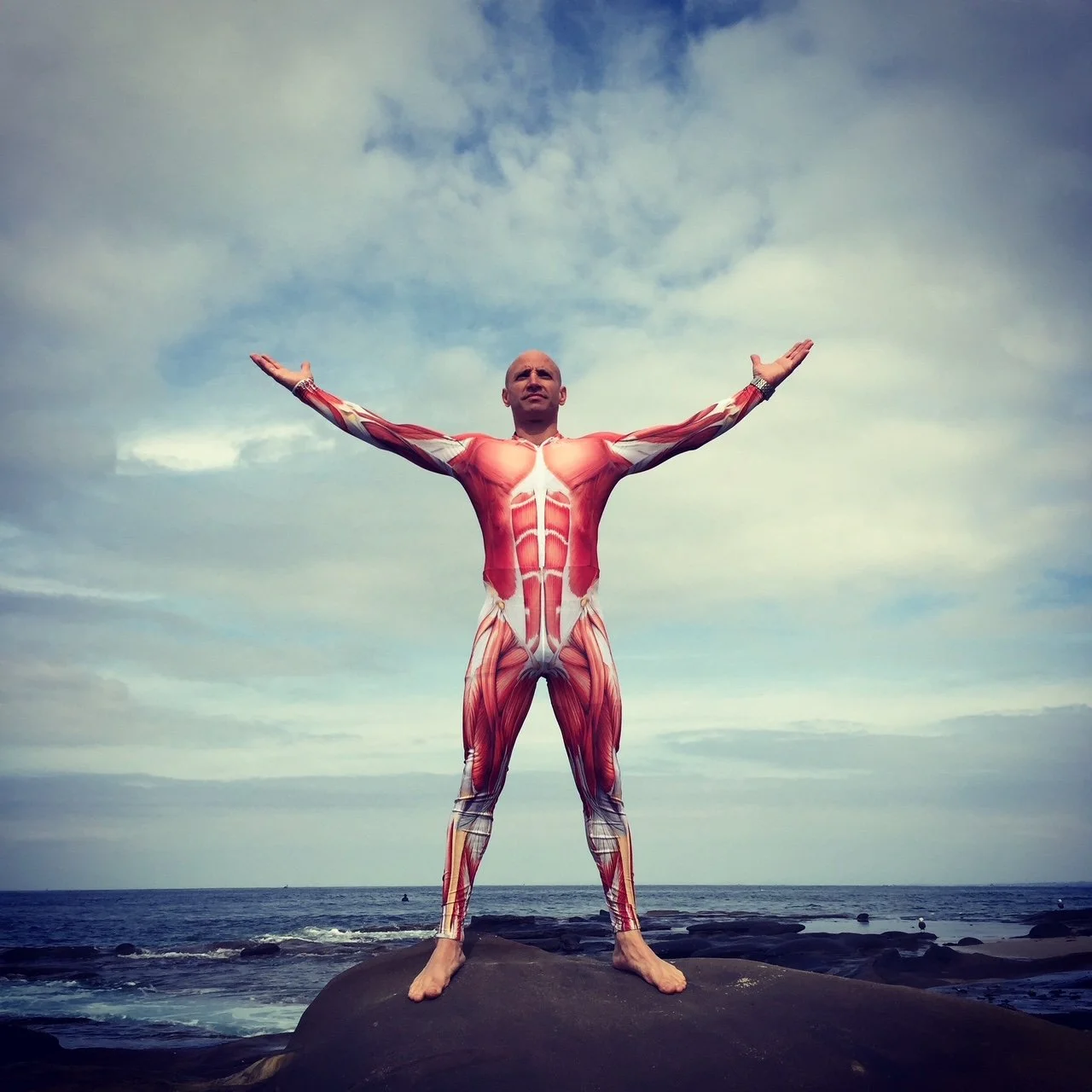 A man standing outdoors on a rock by the ocean with arms outstretched, his body covered with images of human muscle anatomy.