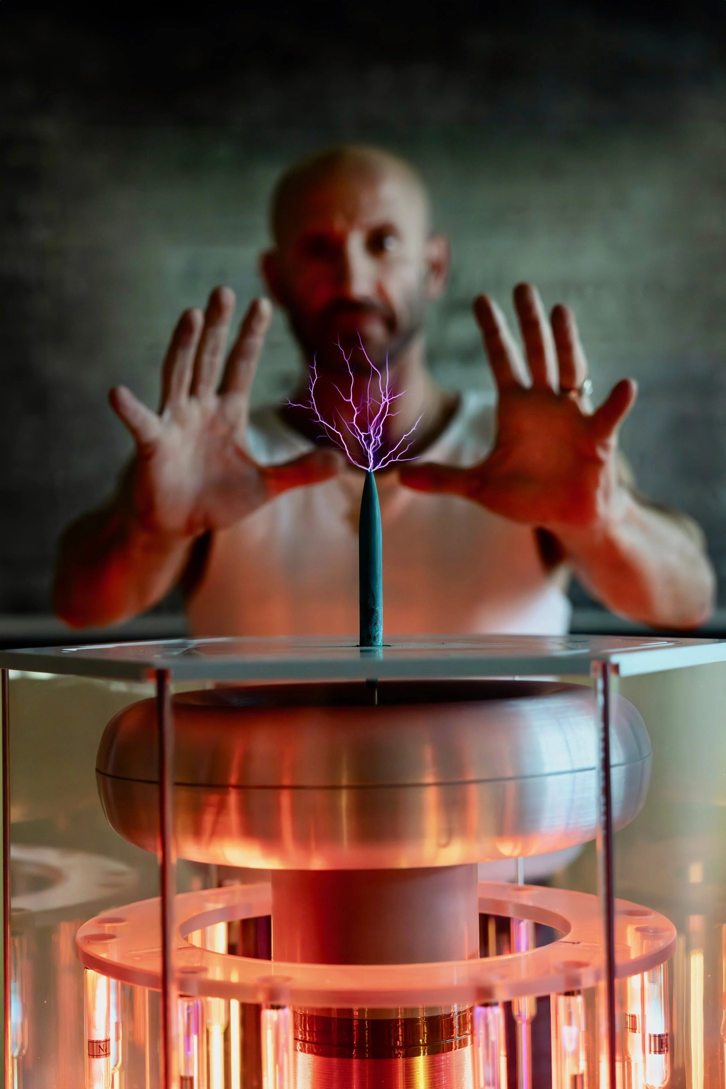A person with a shaved head and beard standing behind a Tesla coil generating purple sparks, with hands raised near the coil, in a dimly lit room.