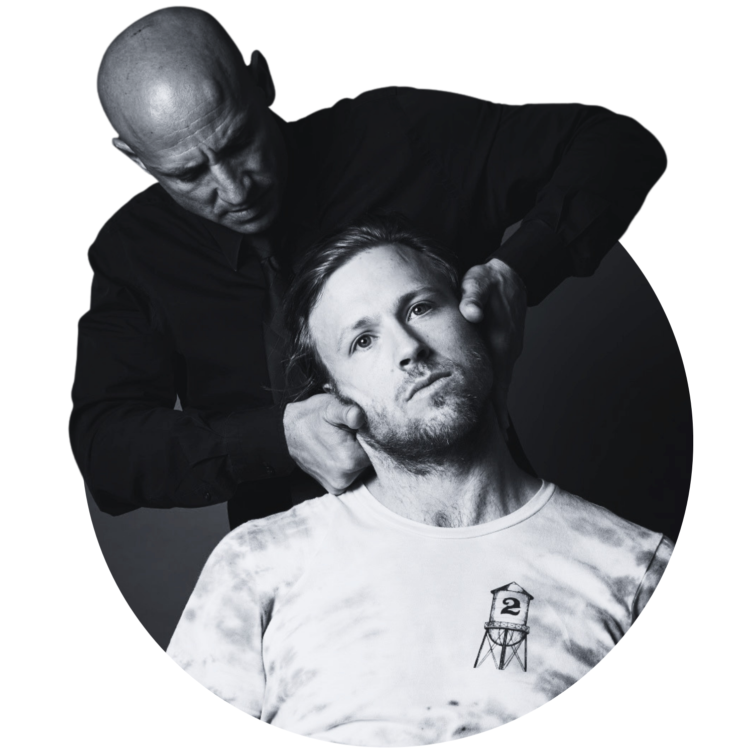 A man with long hair and a beard wearing a graphic T-shirt is having his neck and head examined or massaged by a bald man in black clothing in a monochrome setting.