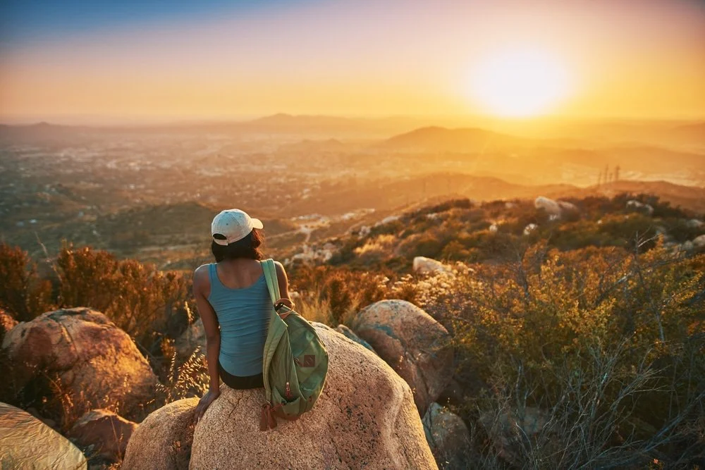 A woman sitting on a rock overlooking a desert landscape during sunset, wearing a cap, tank top, and carrying a green backpack. San Diego, CA