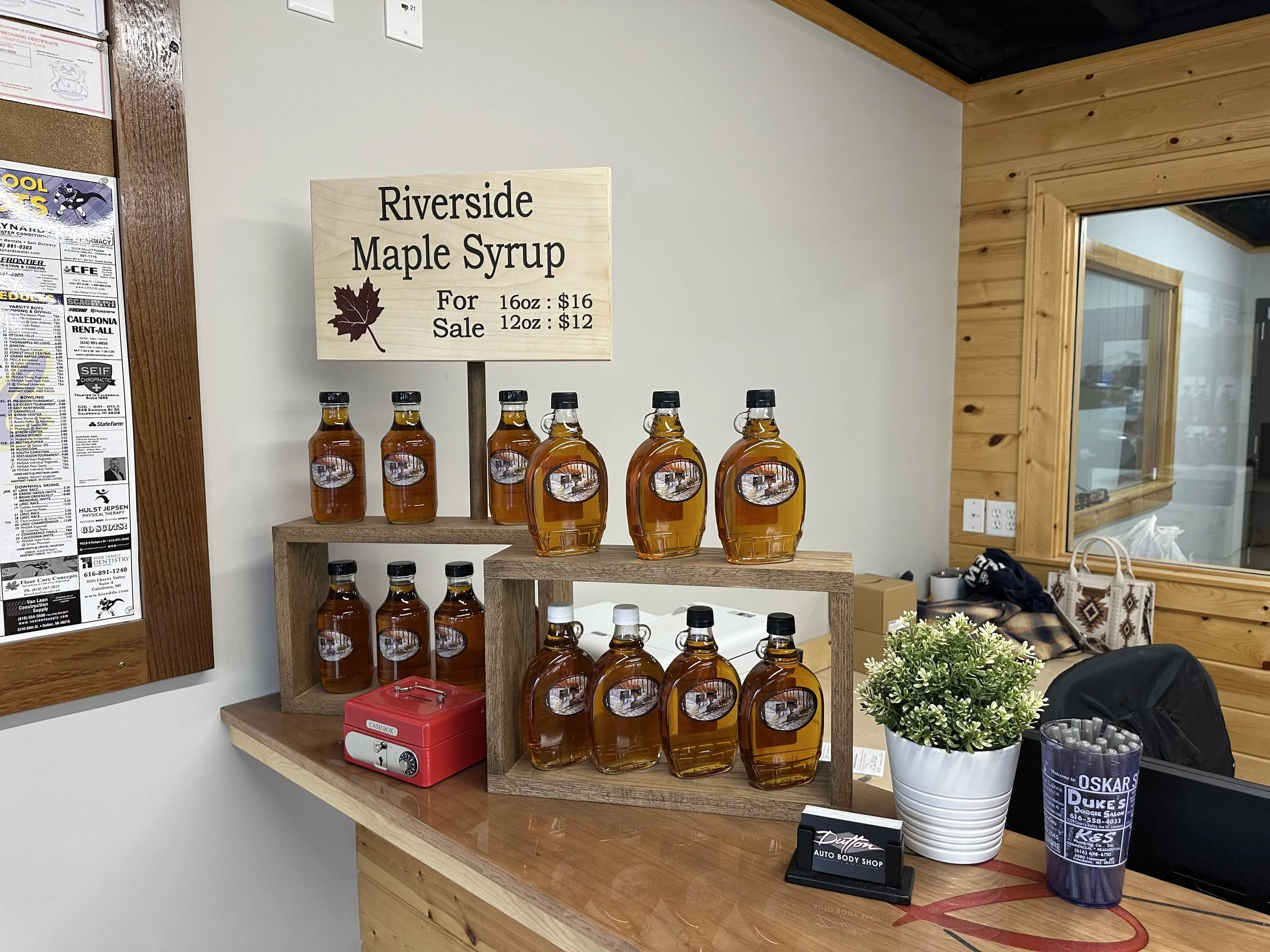 Display of bottles of maple syrup with a sign advertising Riverside Maple Syrup, priced at $16 for 16 ounces and $12 for 12 ounces, on a wooden shelf in an interior space.