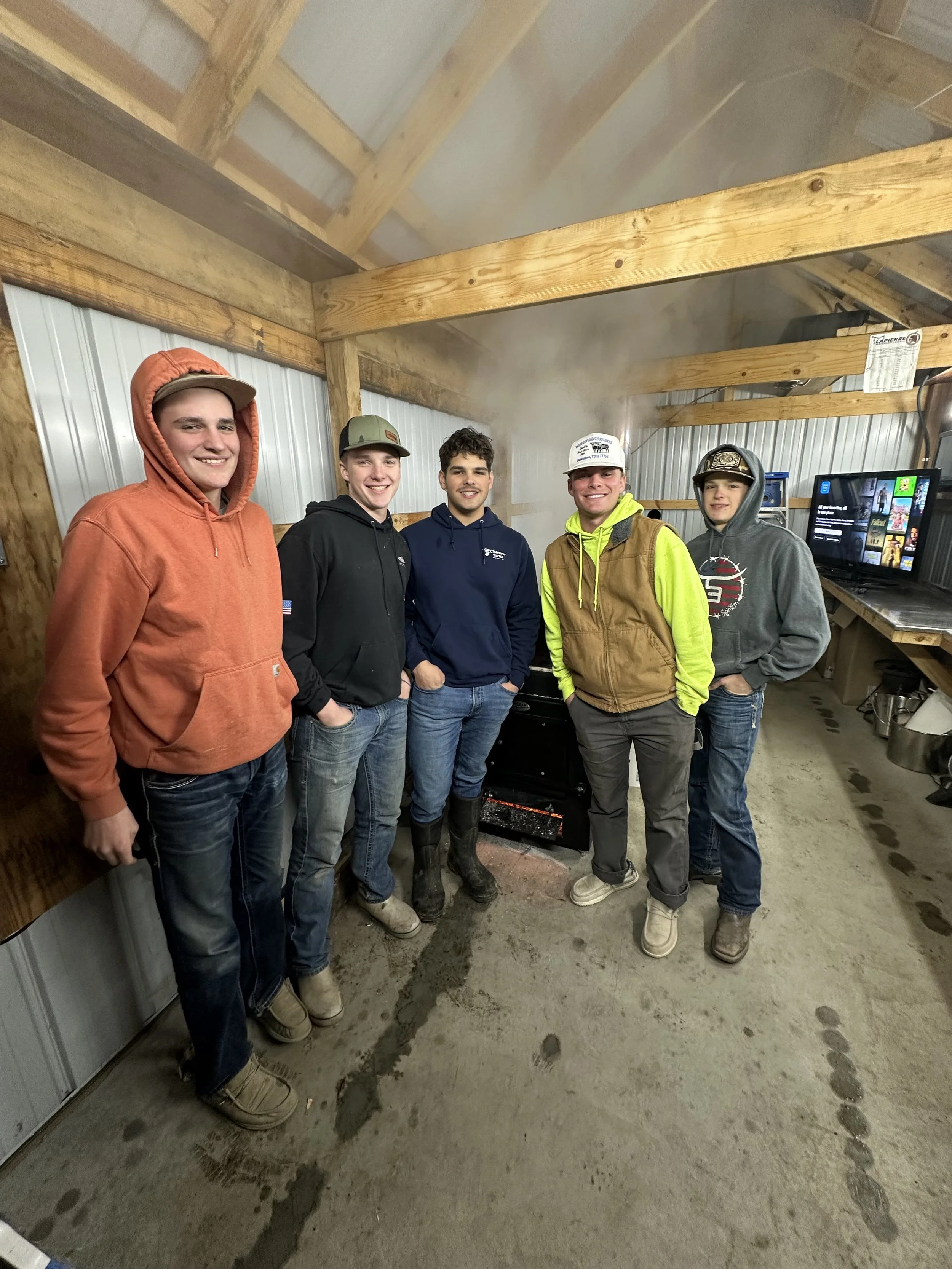 Five young men standing inside a wooden structure, smiling at the camera, dressed in casual winter clothing.