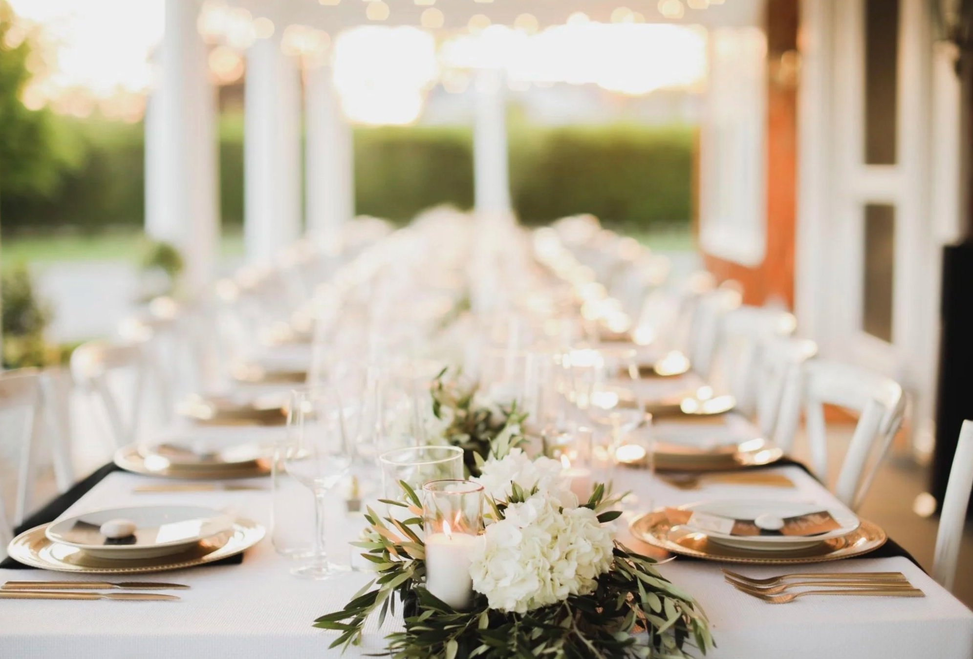 Elegant outdoor dining table set for a wedding or special event, decorated with white flowers, candles, and gold-rimmed plates, with a lush green garden visible in the background.