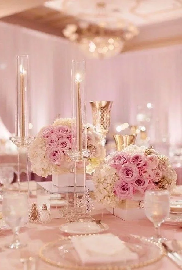 Elegant table setting with pink and white floral centerpieces, tall candles, and gold and crystal accents in a softly lit room.