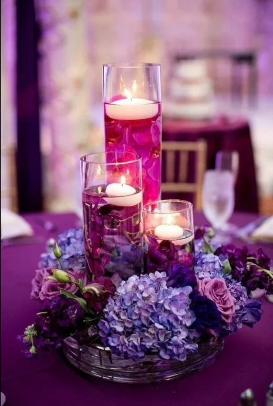 Elegant table centerpiece with three tall glass candle holders containing lit candles, surrounded by purple hydrangeas, roses, and other flowers on a purple tablecloth, in a decorated dining setting.