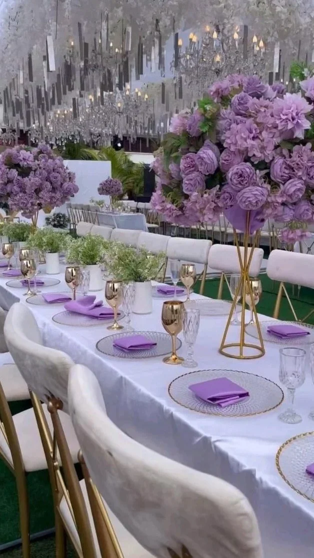 Elegant table setup with white tablecloth, purple napkins, gold and clear glassware, and large purple floral centerpieces. Overhead chandelier with hanging crystals adds a sophisticated ambiance.