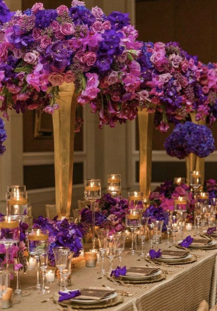 Elegant banquet table decorated with purple and pink floral arrangements, gold candle holders, and lit candles.