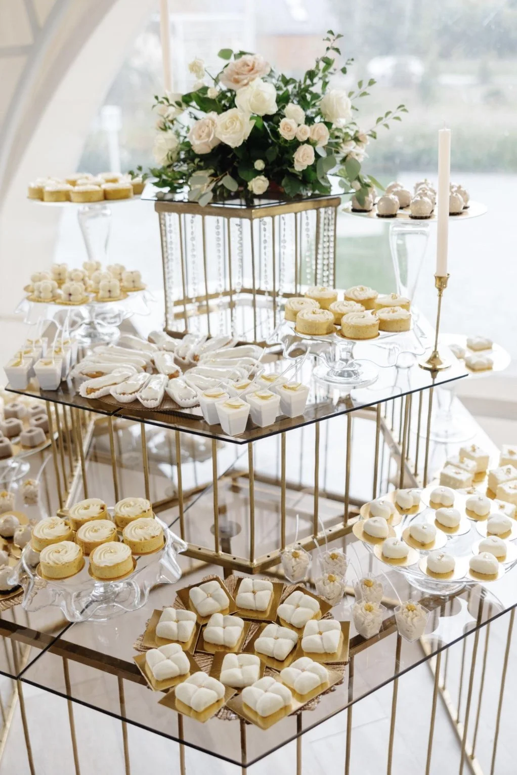 Elegant dessert table with white and cream-colored cookies, cupcakes, and petit fours displayed on glass and gold stands, with a floral centerpiece of white and blush roses and greenery, and a tall white candle in a brass holder.