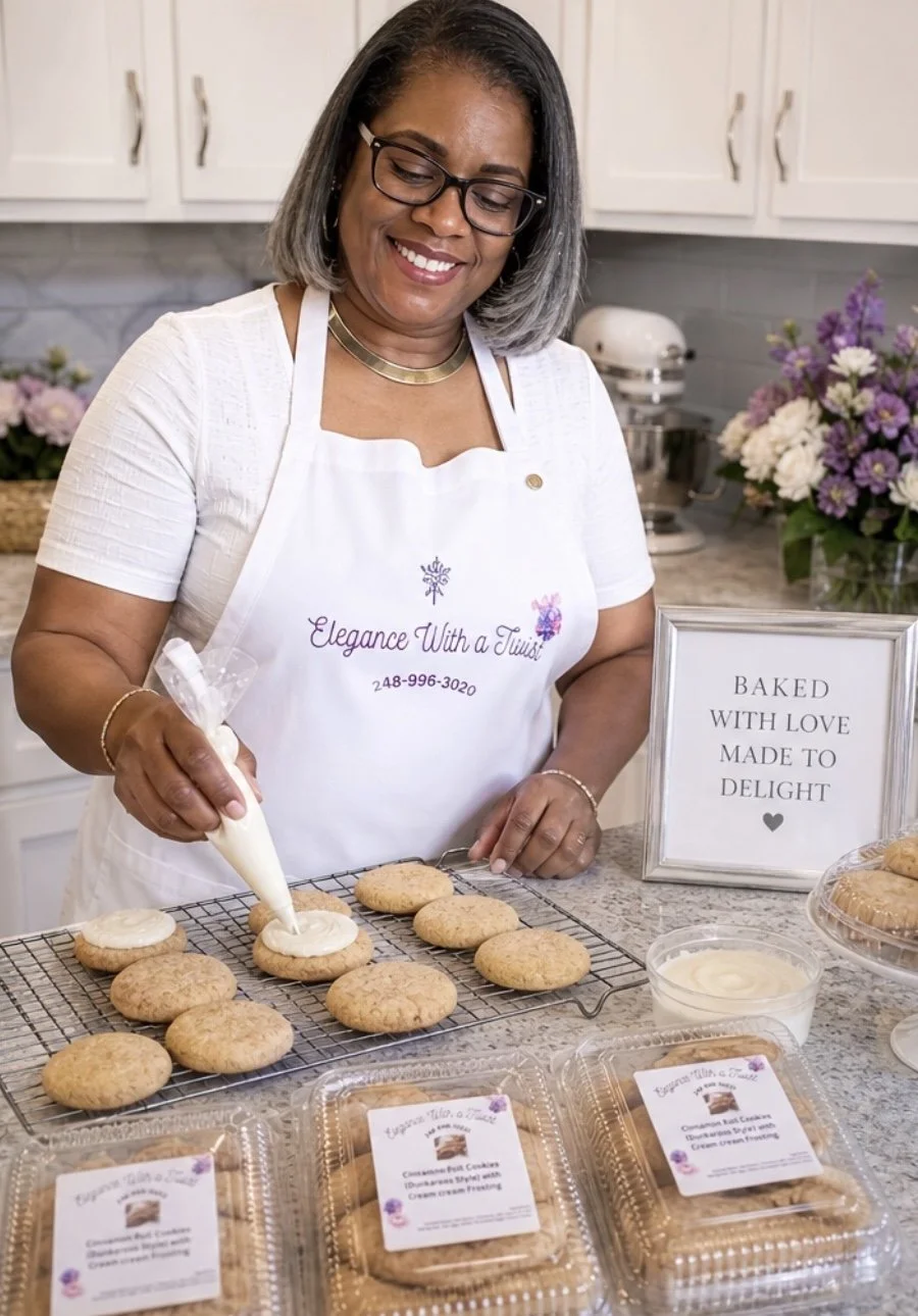 A woman with gray hair and glasses is decorating cookies on a cooling rack in a kitchen. She is wearing a white T-shirt and a white apron with the text 'Elegance With a Twist' and a phone number. There is a framed sign on the counter that says 'Baked with love made to delight,' along with cookies and icing.