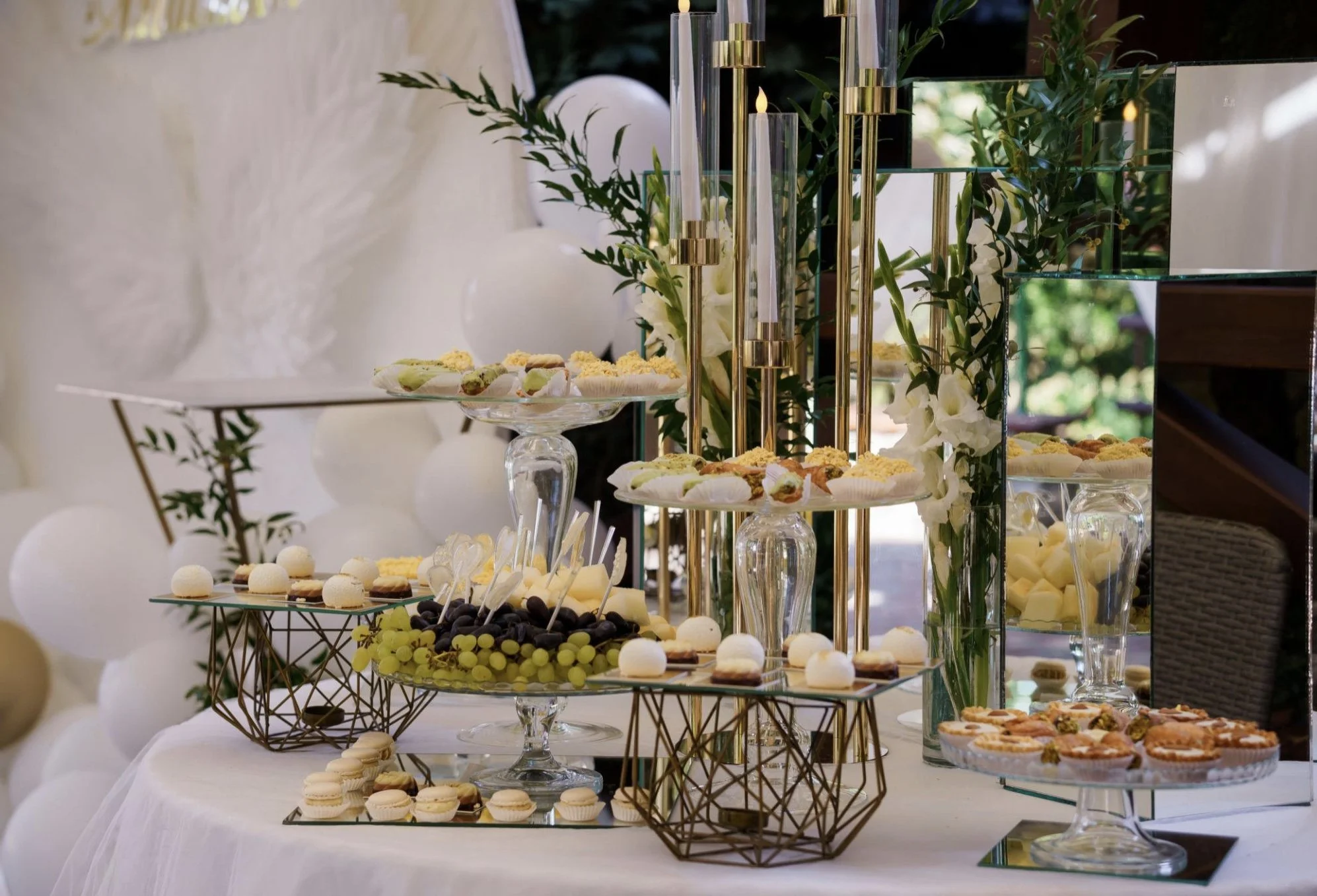 Wedding dessert table with various sweets including macarons, grapes, and mini tarts, decorated with greenery and glass vases, with a backdrop of white balloons and balloons.