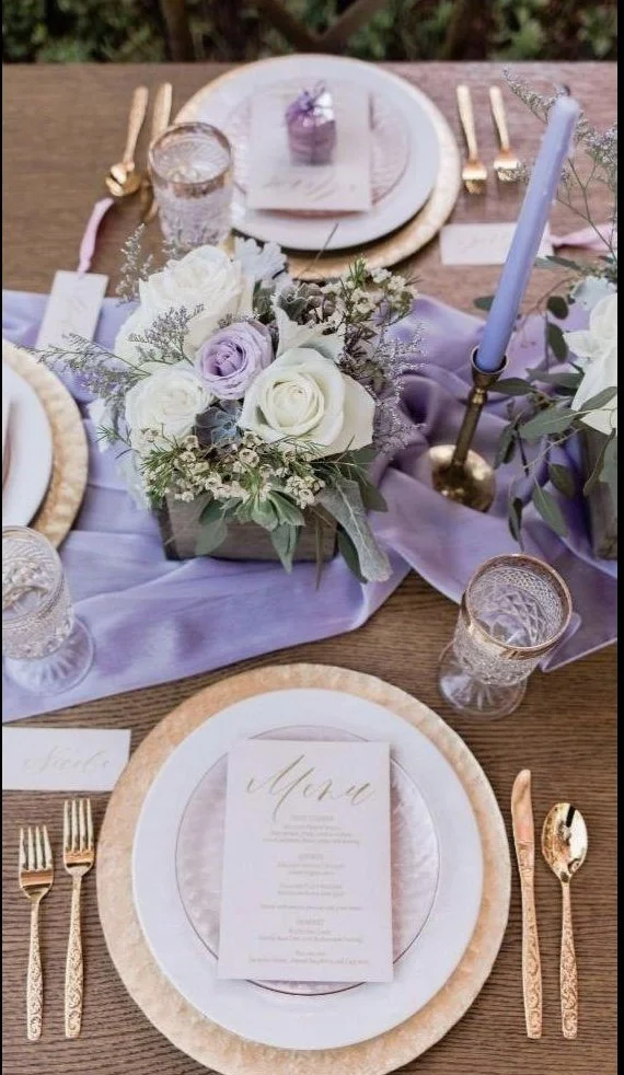 Elegant table setting with a purple-themed floral centerpiece, gold utensils, clear glasses, a menu card, and a tall blue candle in a brass holder, arranged on a purple table runner.