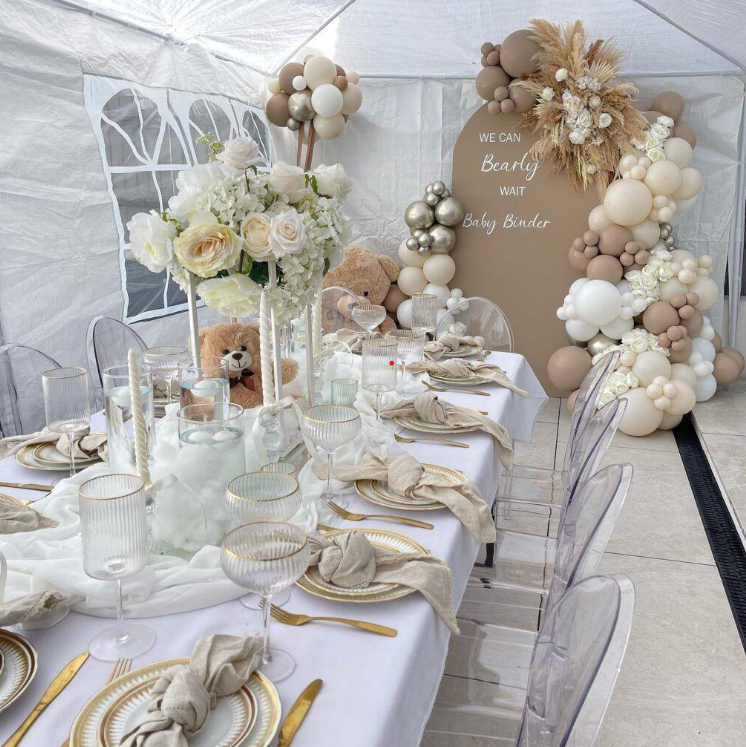 Decorated table at a baby shower with white and beige theme, floral centerpiece, balloon arch, and a sign reading 'We can bearly wait baby bundt.
