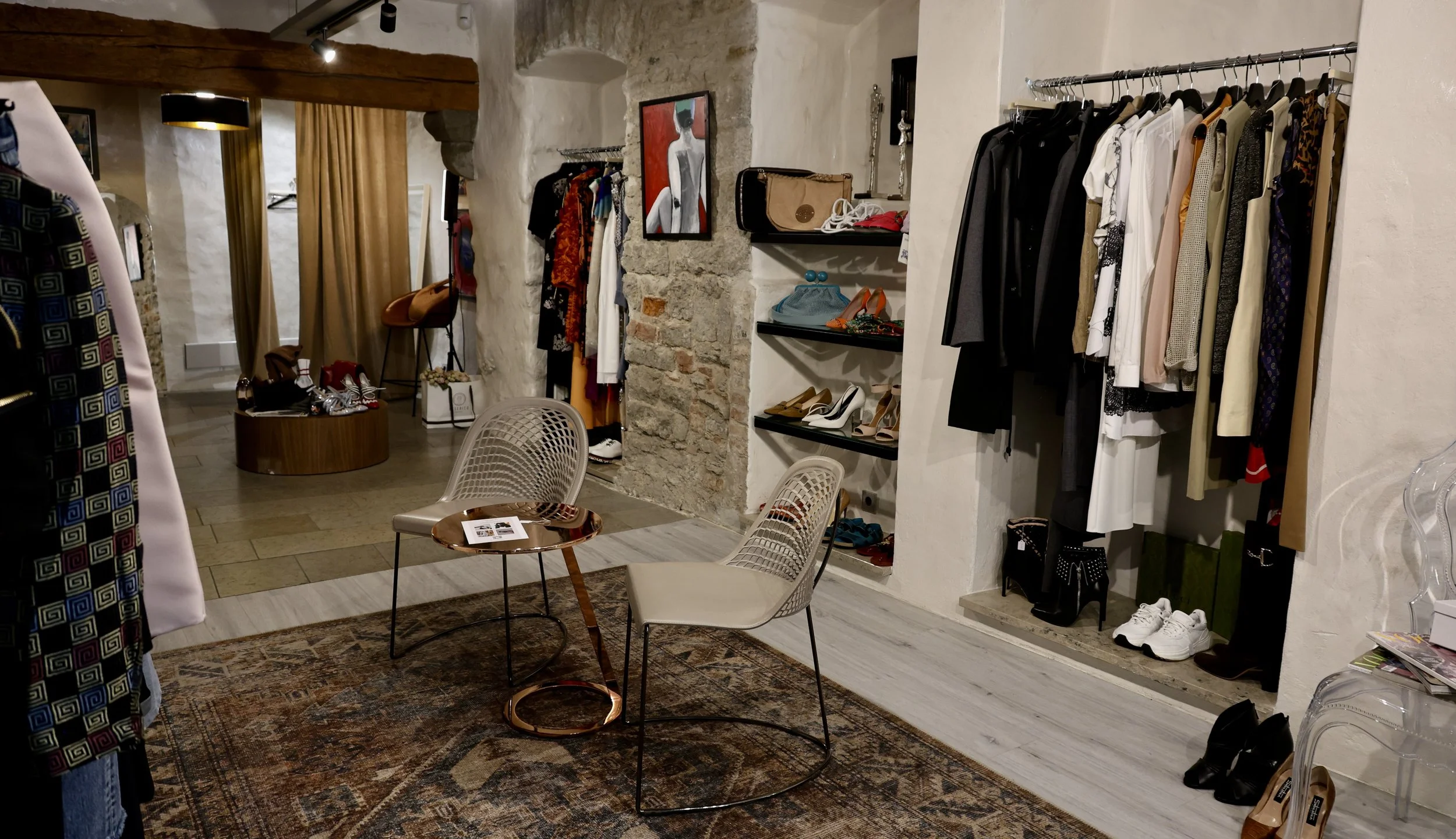A boutique store with clothing racks, shelves with shoes and handbags, a round table with chairs, and a decorative rug. The store has brick and plaster walls with art and accessories displayed.