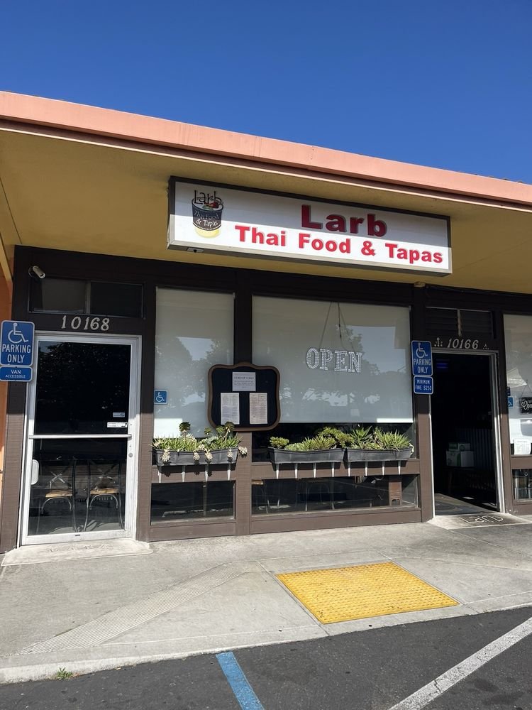 Exterior of a Thai restaurant named Larb with a sign reading 'Thai Food & Tapas', parking signs for handicapped on either side, and a large window with a decorative plant display.