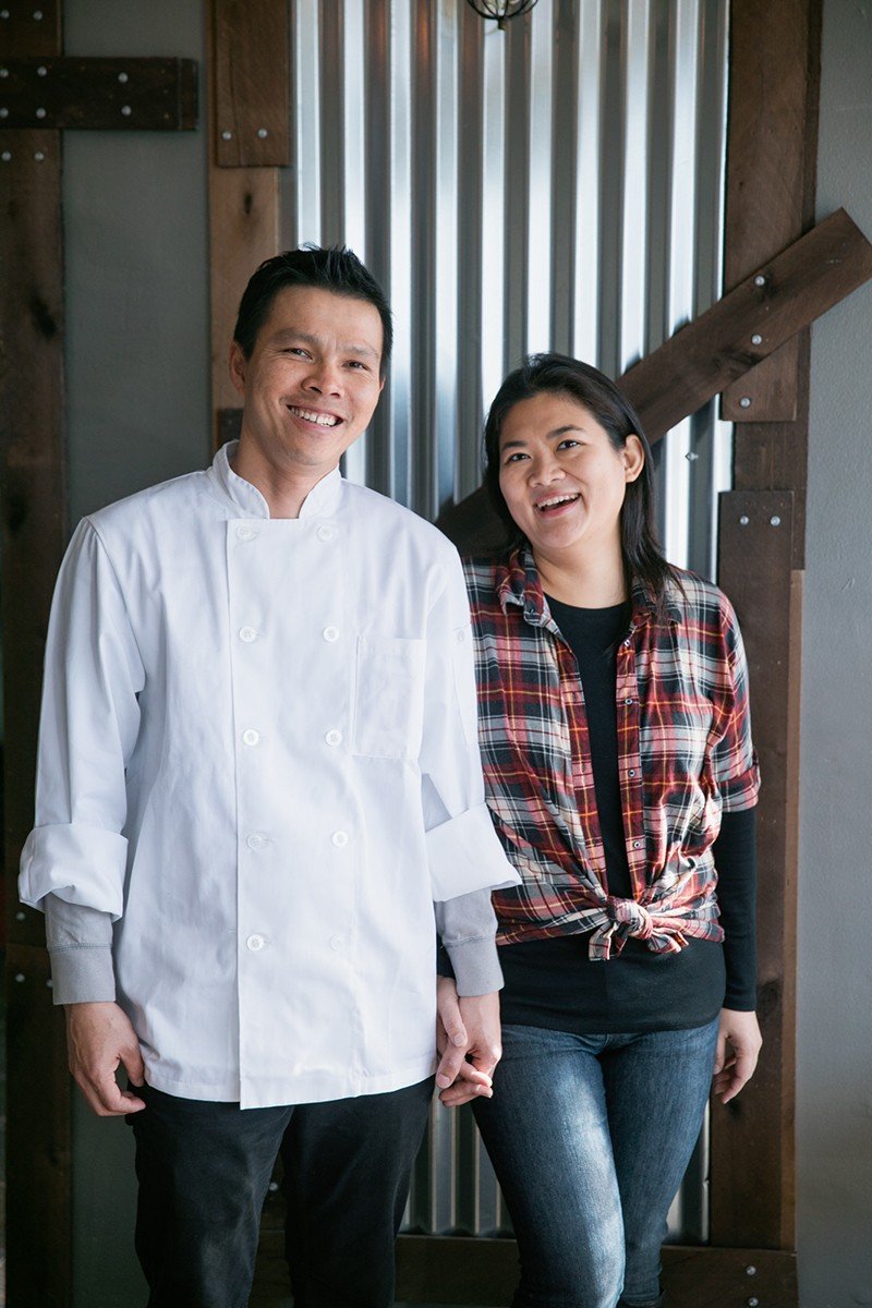 A chef and a woman holding hands and smiling in a rustic kitchen with metal and wooden accents.