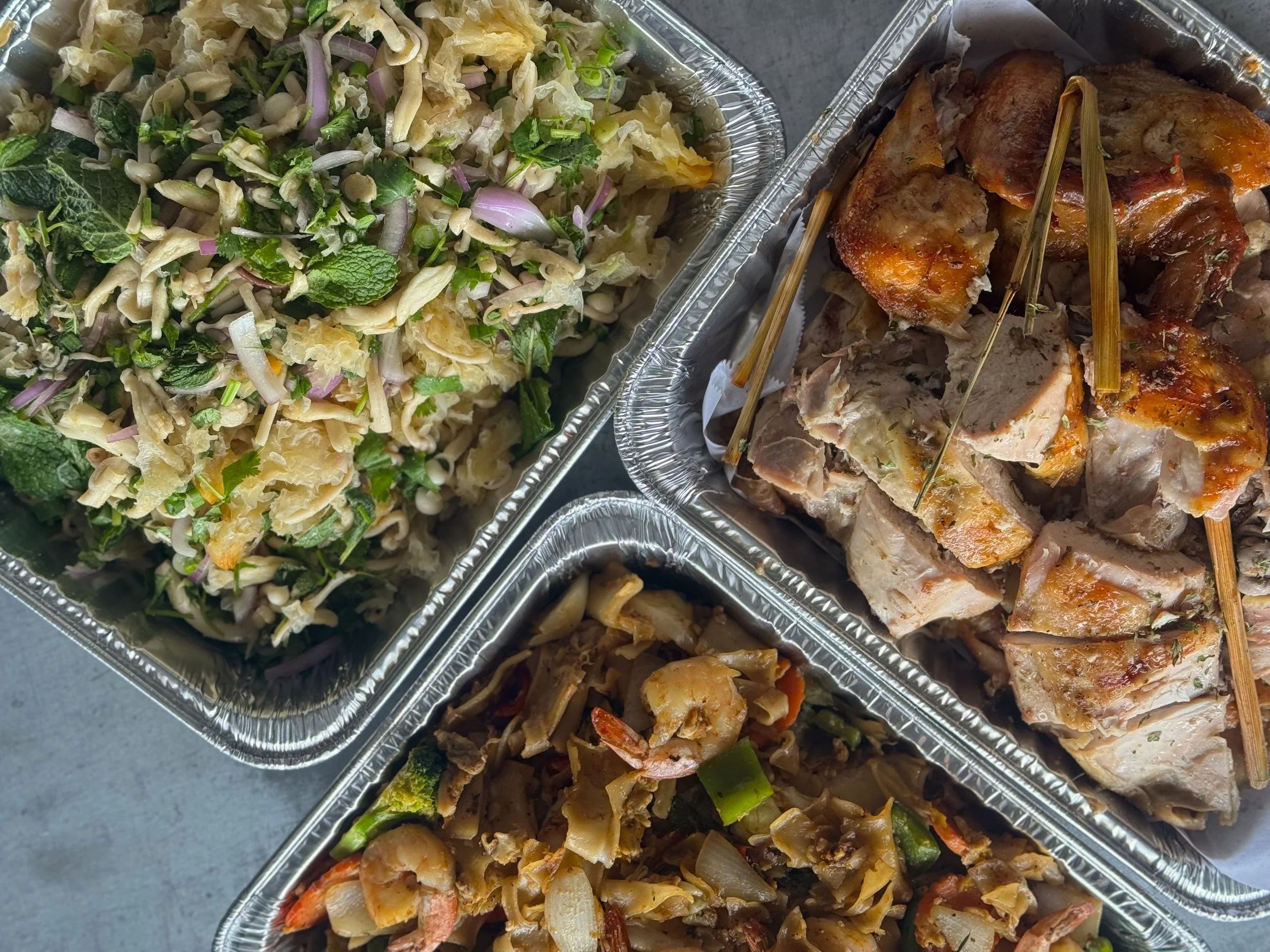 Three aluminum trays of different cooked foods, including a mixed mushroom salad with greens, onions, and herbs on the left, and cooked meat dishes with skewers on the right.
