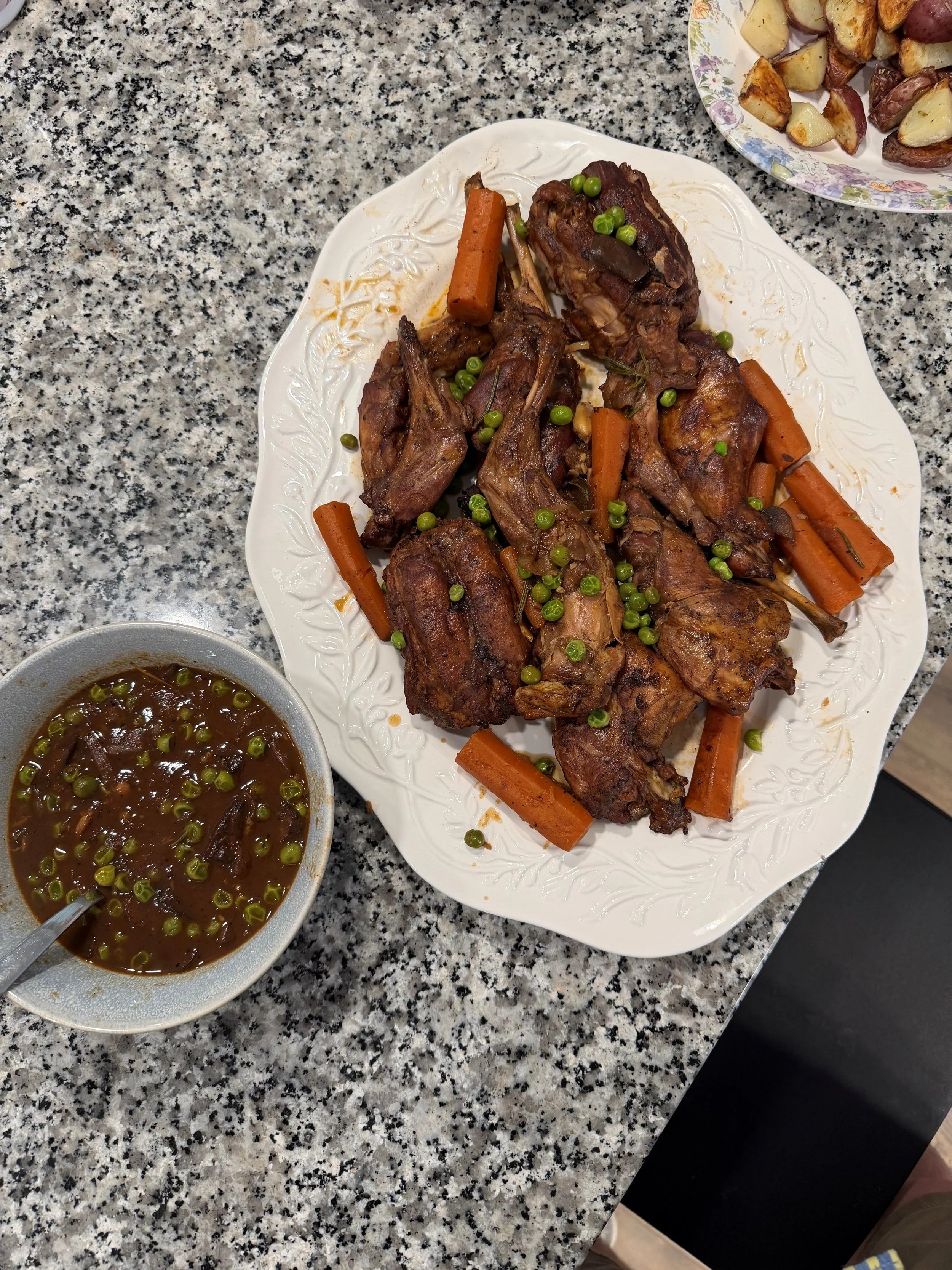 A white plate with cooked lamb chops, carrots, and green peas on a granite countertop, and a bowl of sauce with green peas next to it. In the upper right corner, a bowl of roasted potatoes.