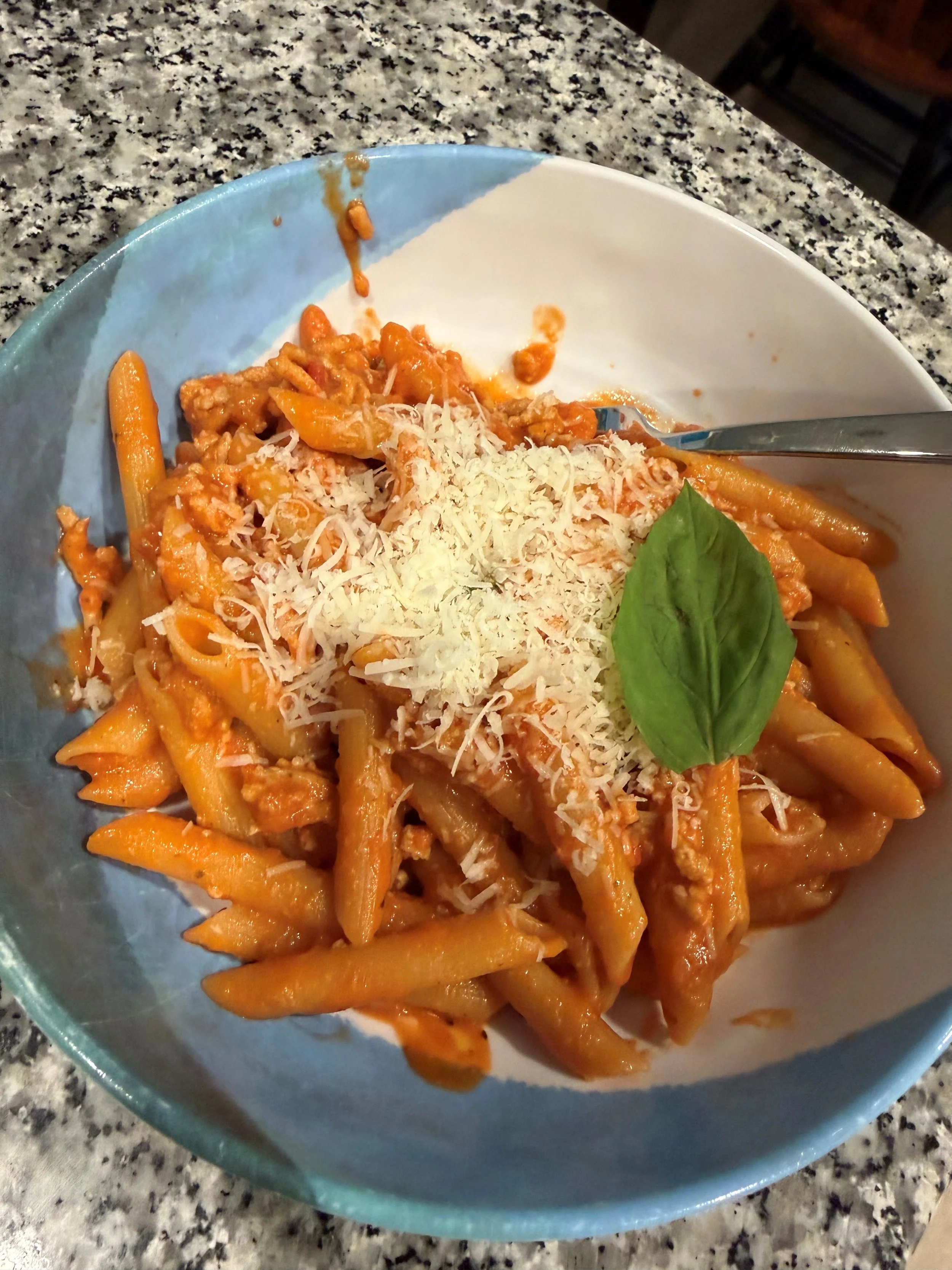 A bowl of cooked penne pasta with orange sauce, grated cheese, and a fresh basil leaf on top, placed on a black and white speckled countertop.
