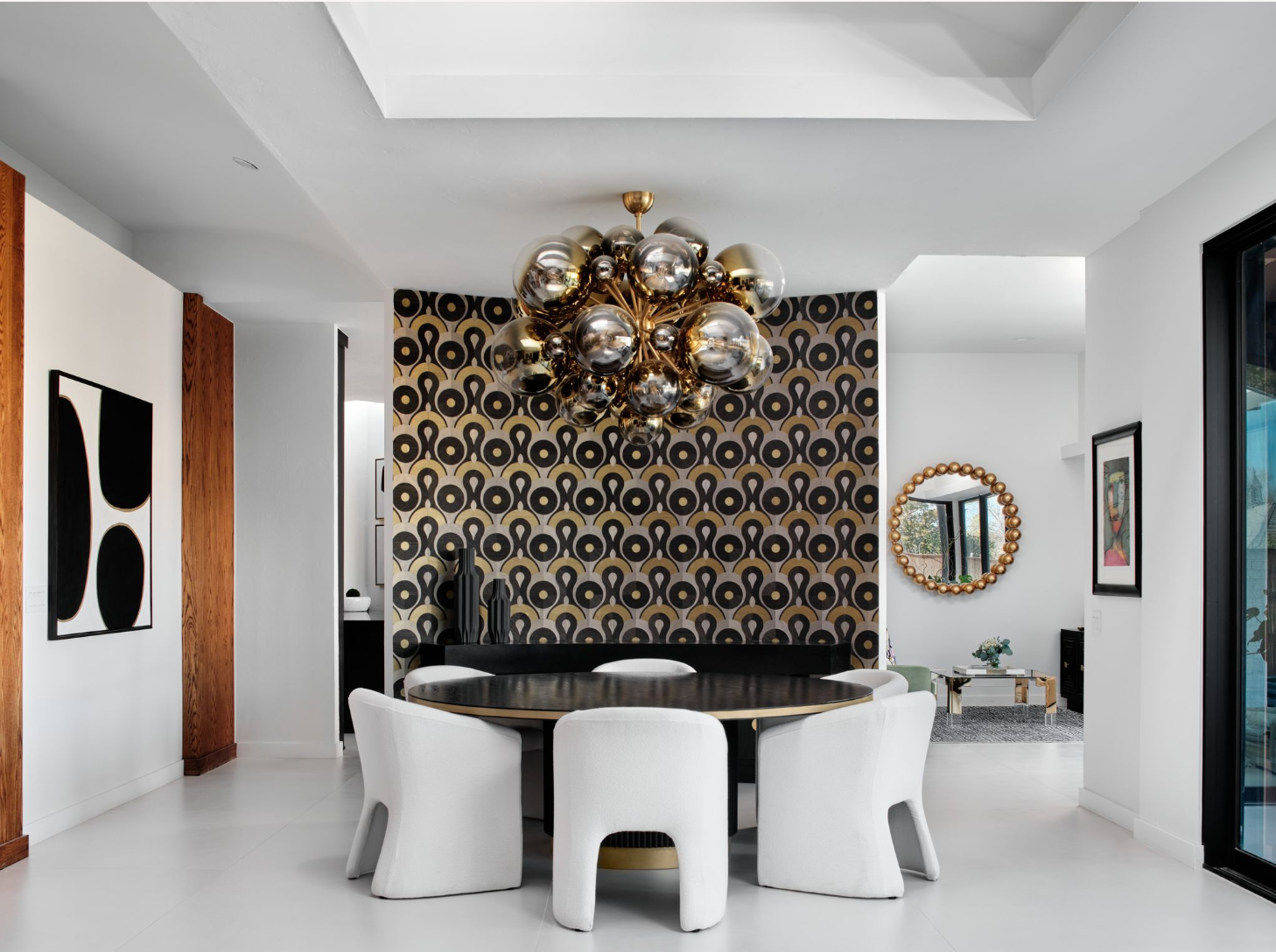 Modern dining room with a black oval table surrounded by six white chairs, a black and gold patterned accent wall, a cluster chandelier with metallic orbs, and decorative black vases on a console table, with artwork and a mirror on the white walls.