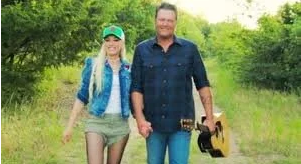 A man and woman walking outdoors in a green, wooded area, holding hands. The woman is wearing a hat, a blue jacket, and khaki shorts, while the man is wearing a dark shirt and jeans, holding a guitar.