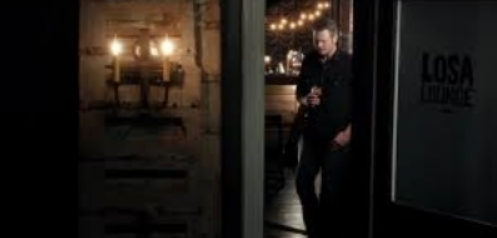 A man standing inside a dimly lit room, looking at his phone, near a door with a sign that reads 'LOSA'.