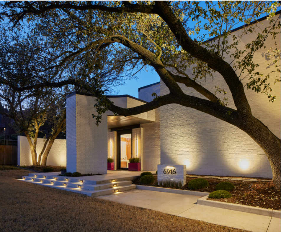 Modern white brick house with entrance illuminated by warm lighting, surrounded by landscaping and large trees at dusk.