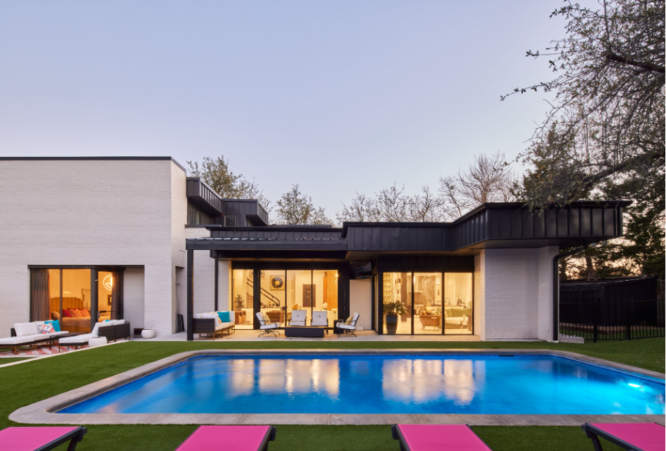 Modern house with large glass windows, outdoor seating area, swimming pool, and pink lounge chairs in the backyard at dusk.