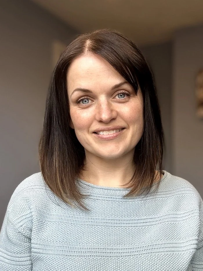 A woman with shoulder-length brown hair, blue eyes, and light skin, smiling indoors against a neutral background.