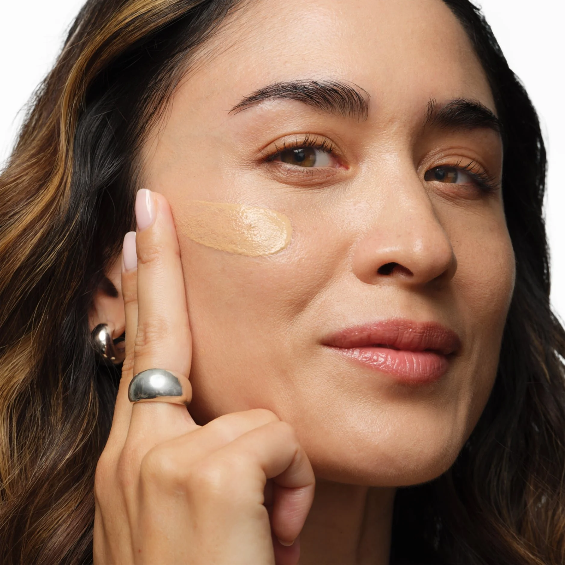 Close-up of a woman with long brown hair applying foundation to her face with her finger.
