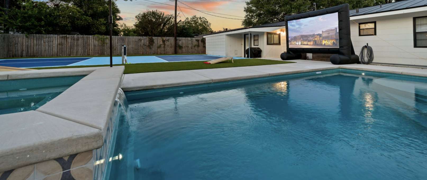 Backyard featuring a swimming pool with a waterfall, a trampoline with a projection screen, and a small building at sunset.