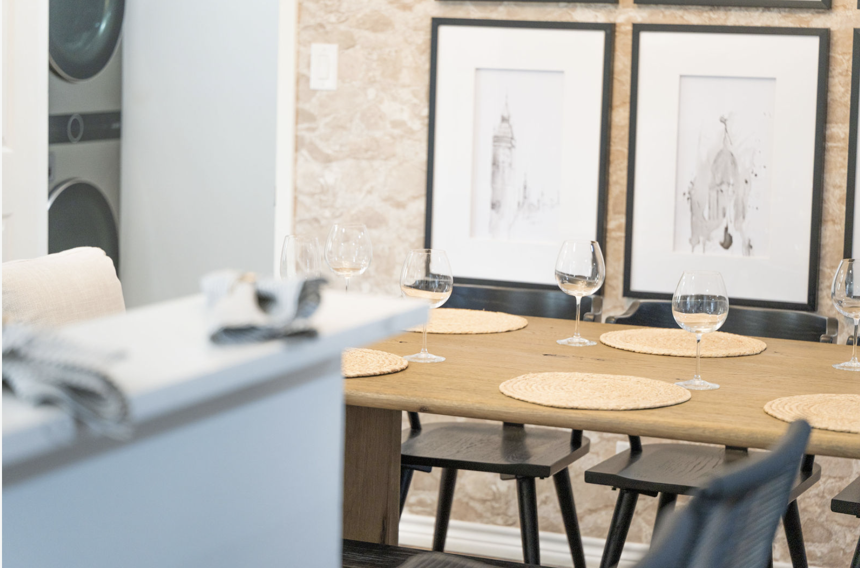 A dining room with a wooden table set with five wine glasses. Four round woven placemats are on the table. There are black chairs around the table. The background features a beige textured wall with two framed art pieces.