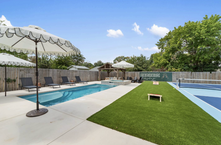 Poolside scene with lounge chairs, umbrellas, a pool, a fenced tennis court, and green trees under a blue sky.