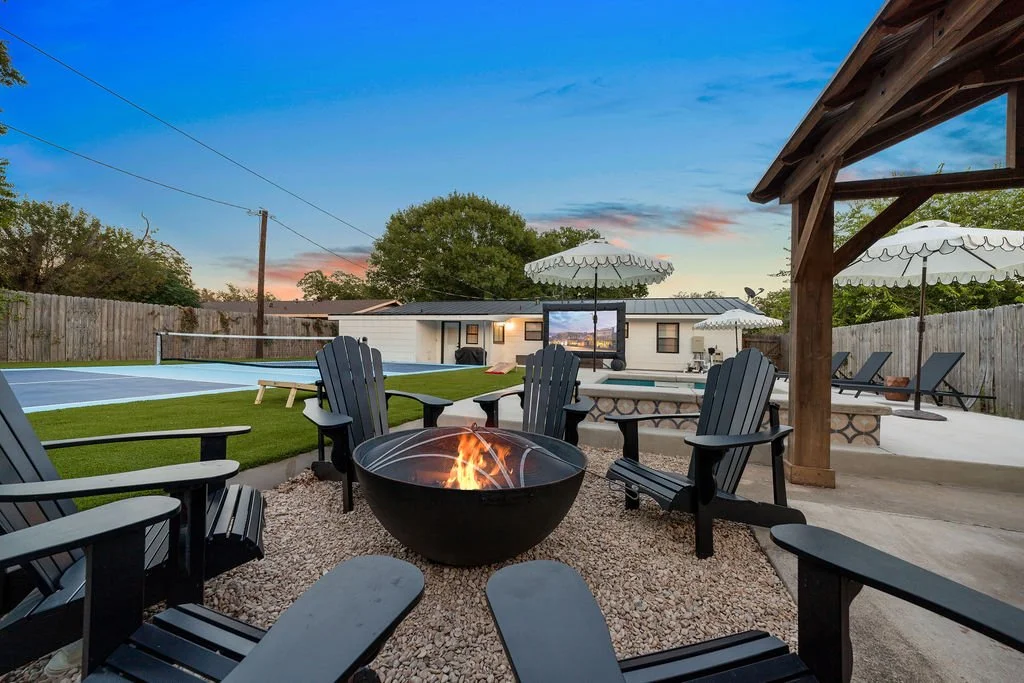 Backyard with a fire pit and Adirondack chairs, a swimming pool with a net, a patio with umbrellas, and a TV, during sunset.