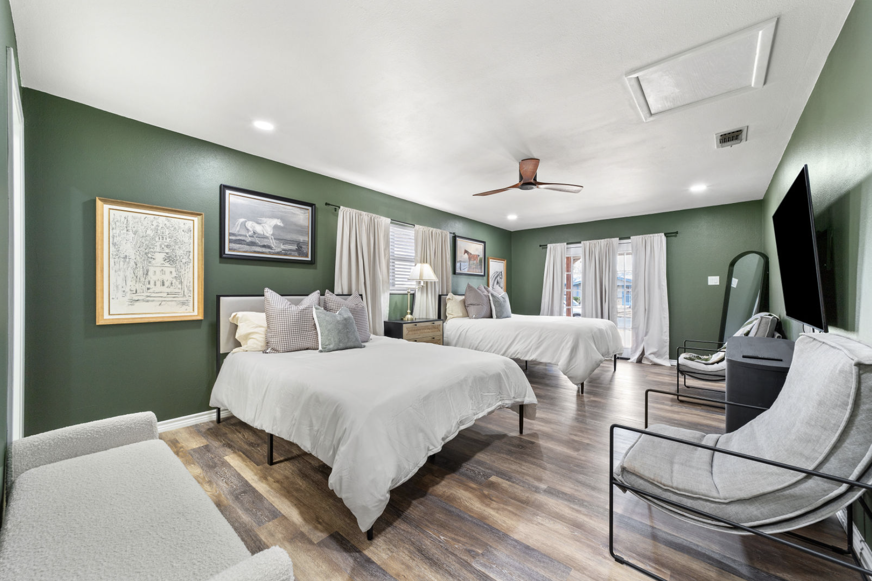 A spacious bedroom with two beds, green walls, framed artwork, and large windows with white curtains. There is a chair with a pillow, a flat-screen TV, and a wooden flooring.