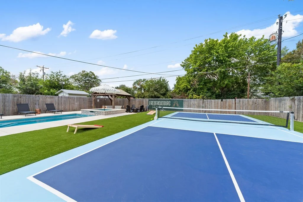 An outdoor pool area with lounge chairs, a gazebo with an umbrella, a ping pong table, and a fenced backyard with trees under a blue sky with clouds.