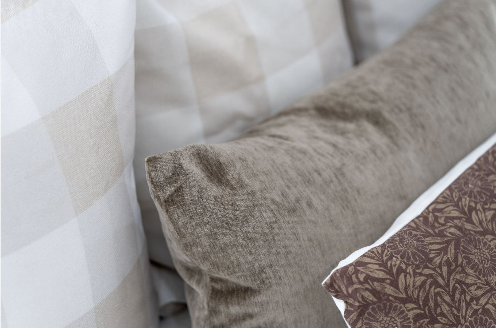 Close-up view of a beige textured pillow on a bed with a patterned brown pillow and a white and beige checkered bedsheet.
