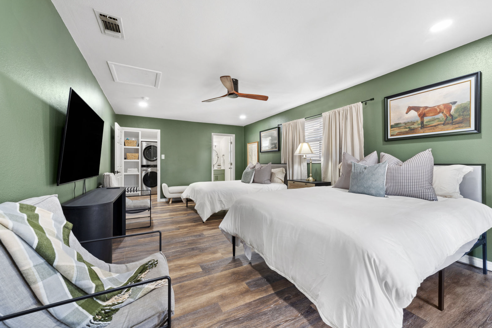 A spacious bedroom with two beds, a black TV on a stand, a green accent wall, and wooden flooring. On the wall above the larger bed is a framed painting of a brown horse. There is a window with curtains, a nightstand with a lamp, and a white chair. A