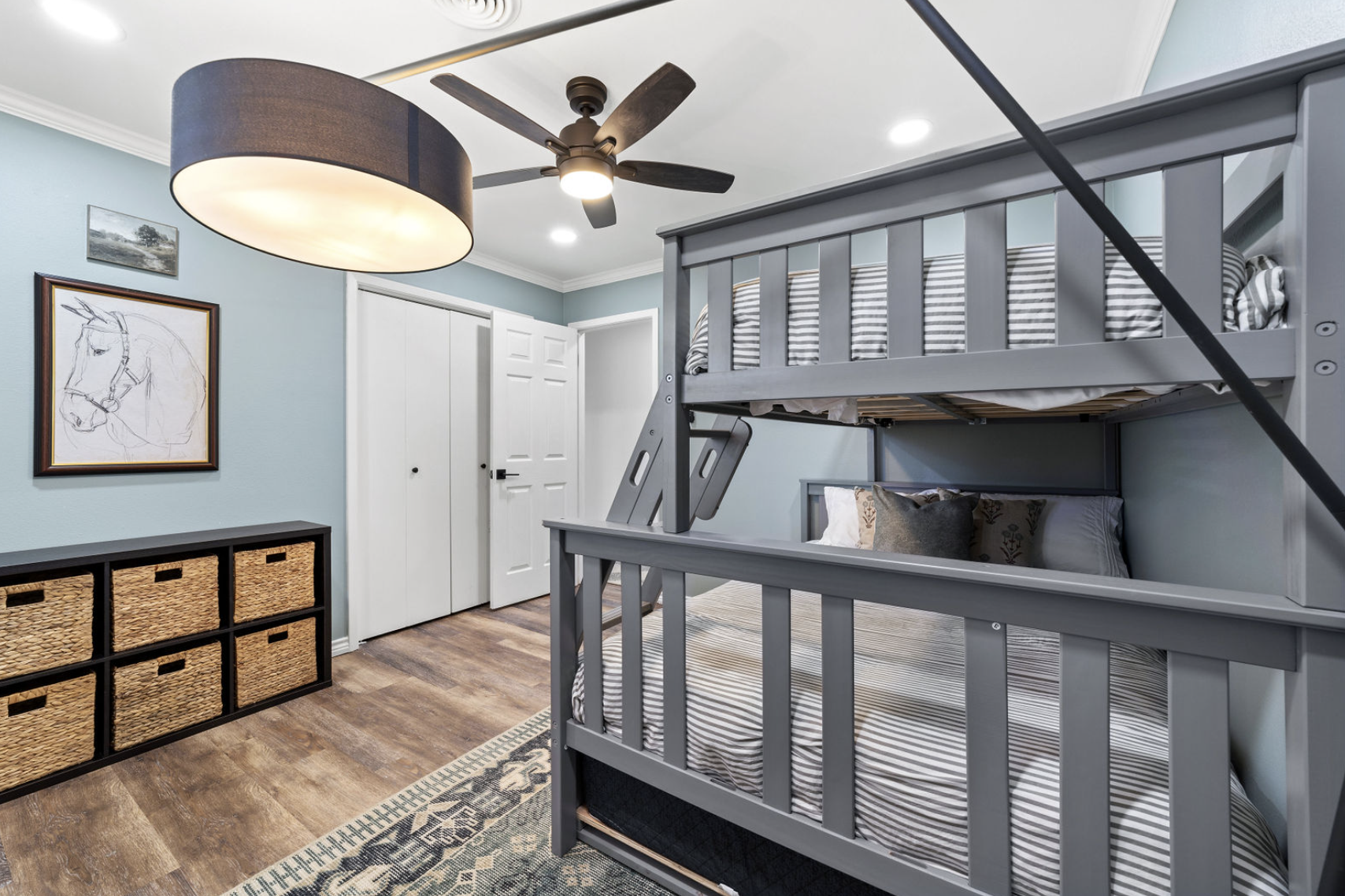 A room with a gray bunk bed, striped bedding, and a gray wall. There's a black storage unit with woven baskets, a framed horse sketch on the wall, a ceiling fan, and a large light fixture.