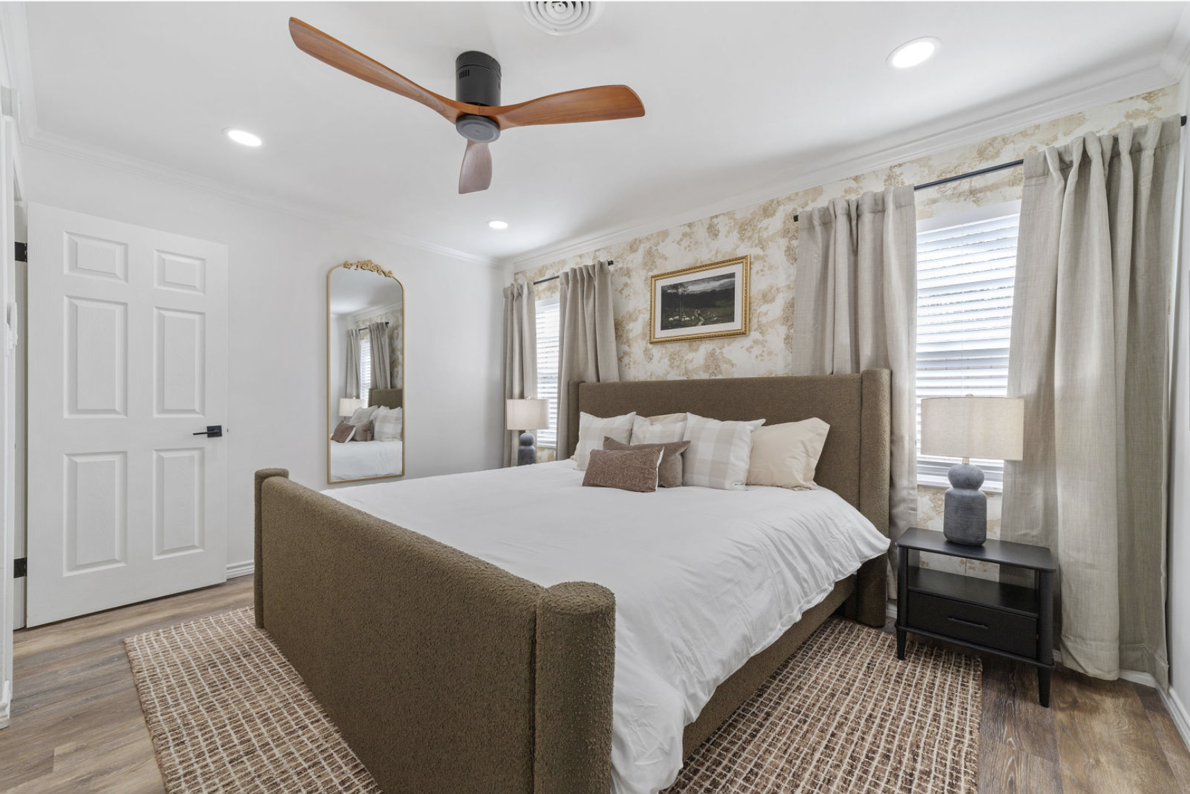 A modern bedroom with a large bed, beige upholstered headboard, white bedding, and multiple pillows. There is a nightstand with a lamp on each side, a wall mirror, and large windows with beige curtains. The walls are decorated with a framed landscape
