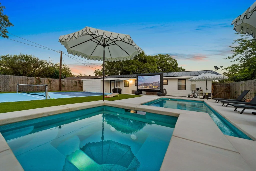 A backyard with a swimming pool and hot tub, outdoor umbrellas, a TV, and lounge chairs, during sunset.