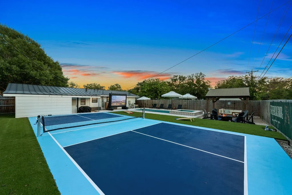 Backyard with a tennis or pickleball court, a swimming pool with umbrellas, a small shed, and outdoor seating at sunset with a colorful sky.