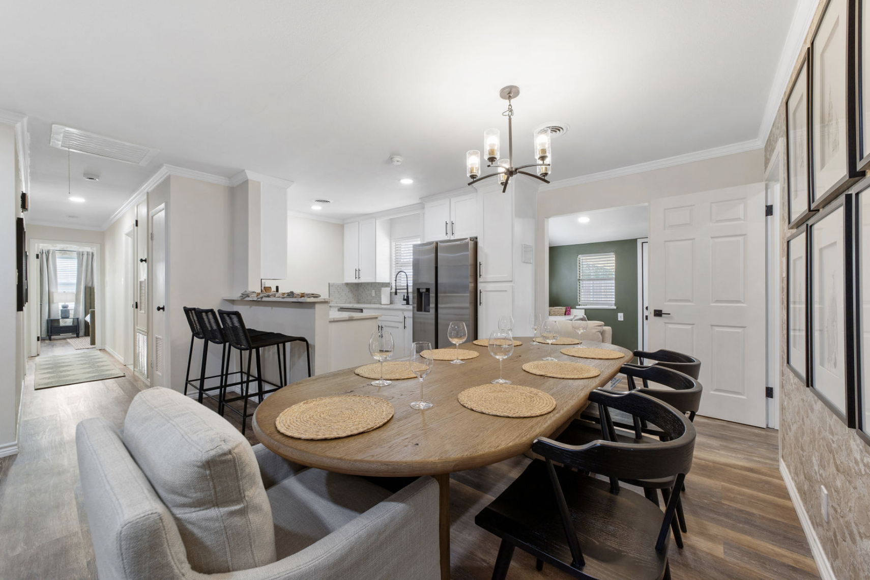 Open-concept kitchen and dining area with a large wooden table set with six placemats and glasses, surrounded by black and beige chairs, white cabinets, stainless steel refrigerator, and decorative wall art.