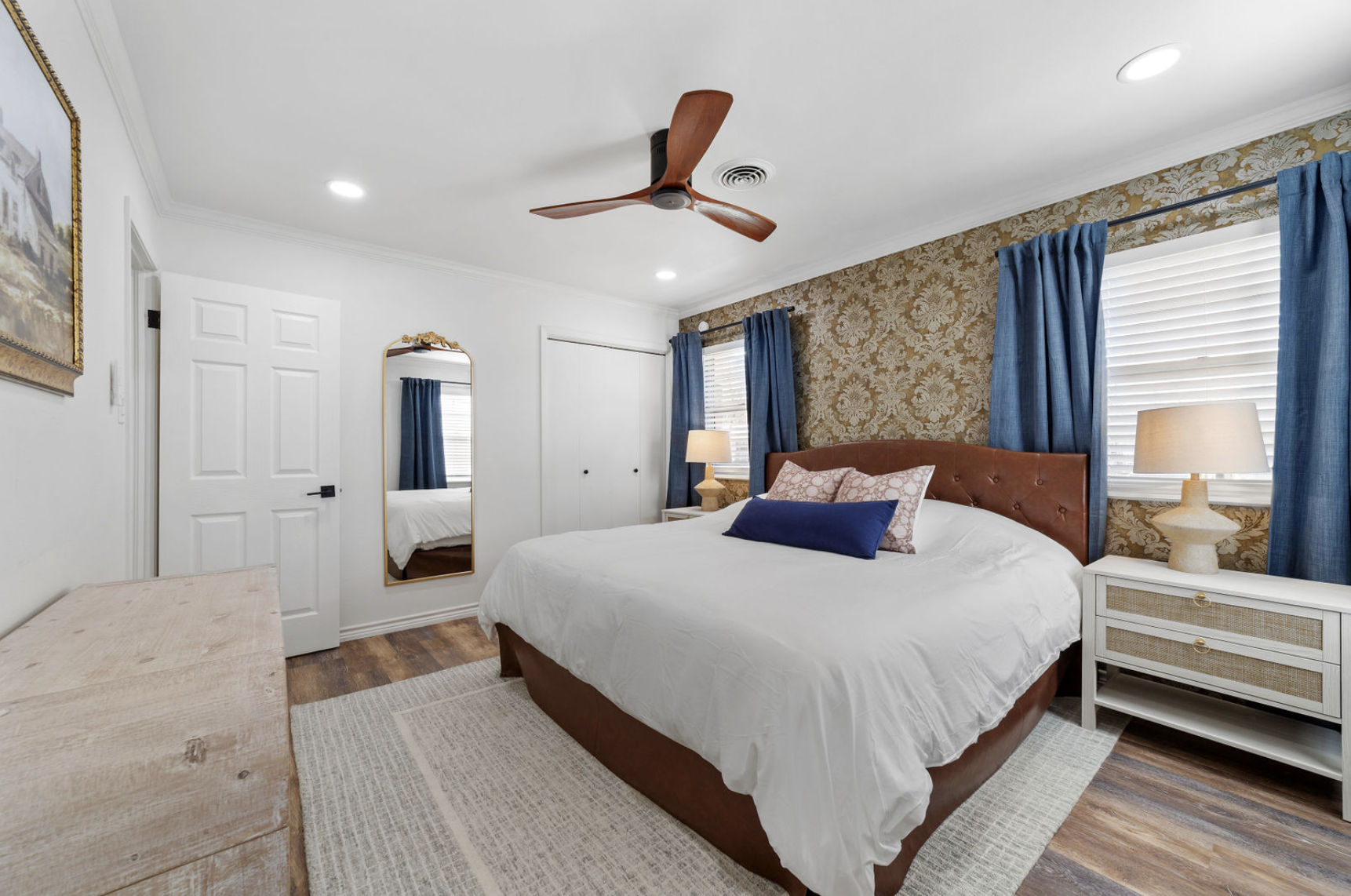 A bedroom featuring a bed with a white quilt, multiple pillows, and a leather headboard. There's a nightstand with a lamp, a large mirror on the wall, blue curtains on two windows, a ceiling fan, and wooden flooring with a beige rug.