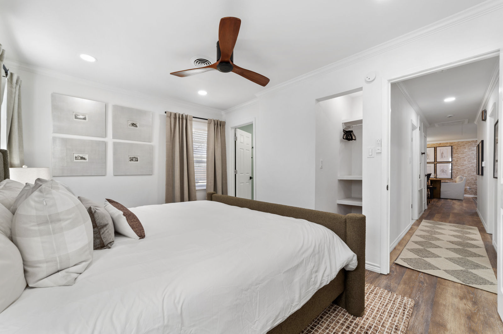 Bedroom with white walls, bed with white bedding and multiple pillows, brown headboard, beige curtains over window, picture frames on wall, ceiling fan, hallway with wood flooring, patterned rug, additional room with pictures on the wall